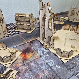 May include: A collection of laser-cut wooden miniature terrain pieces for tabletop gaming, featuring ruined building walls, barricades, and storage containers with etched details, arranged on a printed urban combat game mat.