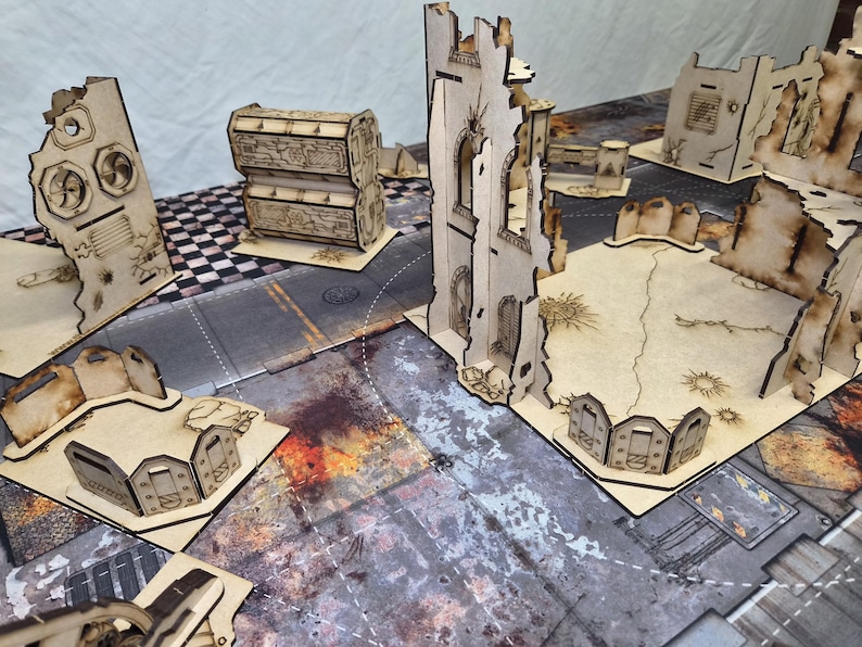 May include: A collection of laser-cut wooden miniature terrain pieces for tabletop gaming, featuring ruined building walls, barricades, and storage containers with etched details, arranged on a printed urban combat game mat.