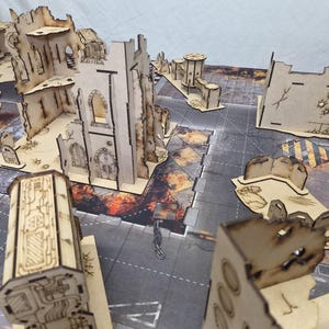 May include: A collection of laser-cut wooden terrain pieces for tabletop gaming, featuring ruined building structures with scorched edges, gothic-style arches, and industrial wall segments arranged on a printed urban battle mat surface.