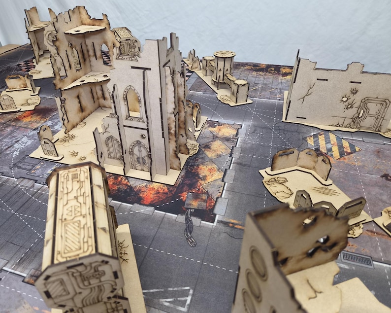 May include: A collection of laser-cut wooden terrain pieces for tabletop gaming, featuring ruined building structures with scorched edges, gothic-style arches, and industrial wall segments arranged on a printed urban battle mat surface.