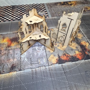 May include: Two beige laser-cut wooden structures representing ruined buildings with arched windows and jagged edges, positioned on a printed gaming mat featuring a gray urban street grid with patches of fire and debris patterns.