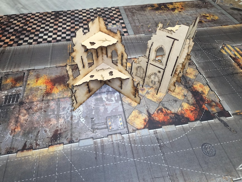 May include: Two beige laser-cut wooden structures representing ruined buildings with arched windows and jagged edges, positioned on a printed gaming mat featuring a gray urban street grid with patches of fire and debris patterns.
