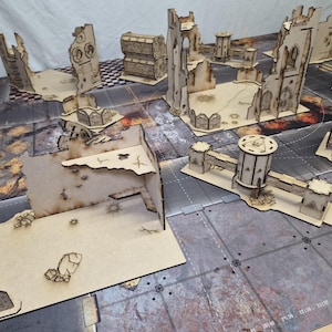 May include: A collection of laser-cut wooden tabletop wargaming terrain pieces featuring ruined building walls, defensive bunkers, and cylindrical towers with scorched edges, arranged on a printed game mat with a grid and industrial textures.