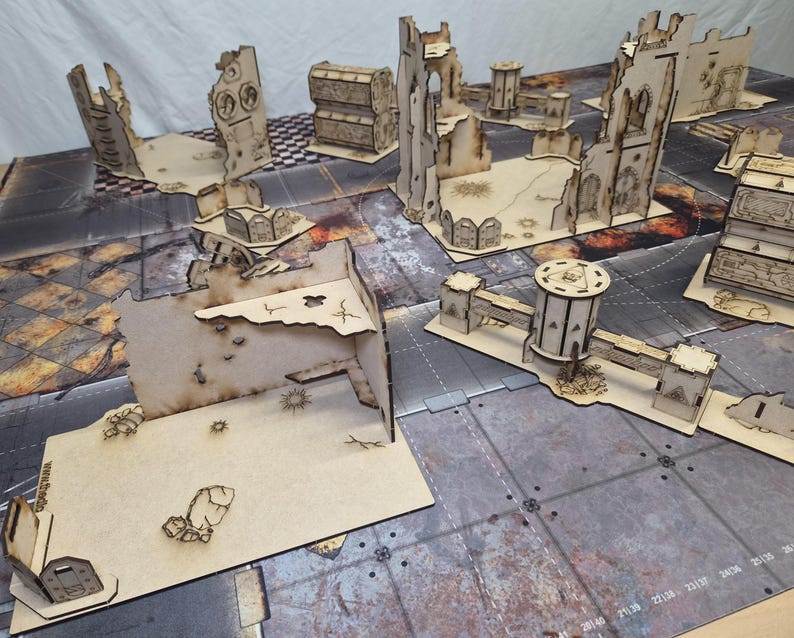 May include: A collection of laser-cut wooden tabletop wargaming terrain pieces featuring ruined building walls, defensive bunkers, and cylindrical towers with scorched edges, arranged on a printed game mat with a grid and industrial textures.