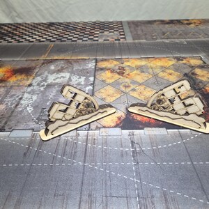 May include: Two light-colored laser-cut wooden terrain pieces featuring a jagged, ruined wall design with exposed gear-like mechanical details. These miniature structures are crafted from flat wood panels and stand upright on a textured base.