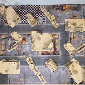 May include: A tabletop gaming terrain set featuring multiple laser-cut tan wooden structures with ruined wall designs, arranged on a printed game mat with a gray urban street layout, checkered patterns, and weathered industrial textures.