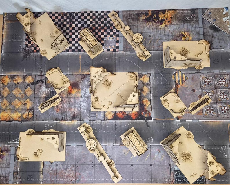May include: A tabletop gaming terrain set featuring multiple laser-cut tan wooden structures with ruined wall designs, arranged on a printed game mat with a gray urban street layout, checkered patterns, and weathered industrial textures.