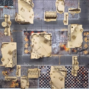 May include: A top-down view of a tabletop gaming mat featuring a printed urban ruin design with gray concrete, checkered, and fire-damaged patterns. Several light-colored laser-cut wooden building ruins are arranged across the surface.