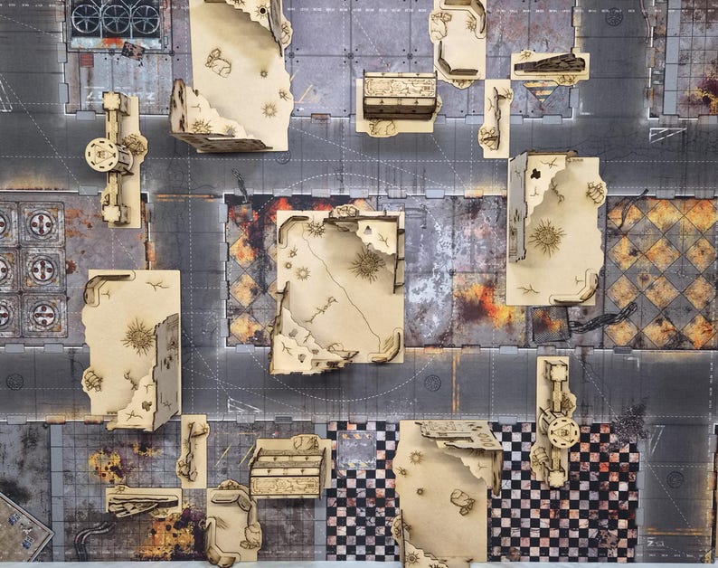 May include: A top-down view of a tabletop gaming mat featuring a printed urban ruin design with gray concrete, checkered, and fire-damaged patterns. Several light-colored laser-cut wooden building ruins are arranged across the surface.