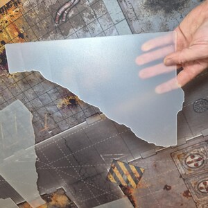 May include: A translucent, frosted plastic sheet with an irregular, jagged edge. The material is semi-opaque and smooth, designed for use as a gaming terrain overlay or template. It features a flat surface and a distinct, non-uniform cut shape.
