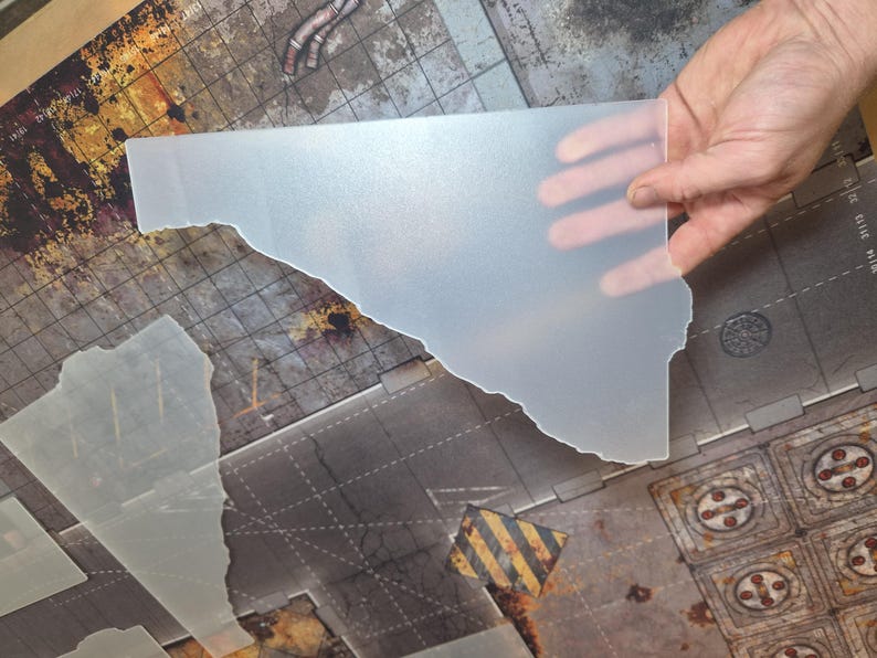 May include: A translucent, frosted plastic sheet with an irregular, jagged edge. The material is semi-opaque and smooth, designed for use as a gaming terrain overlay or template. It features a flat surface and a distinct, non-uniform cut shape.