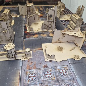 May include: A collection of laser-cut wooden tabletop wargaming terrain pieces arranged on a printed game mat. The set includes various ruined building structures, walls, and defensive barricades featuring etched details and weathered textures.