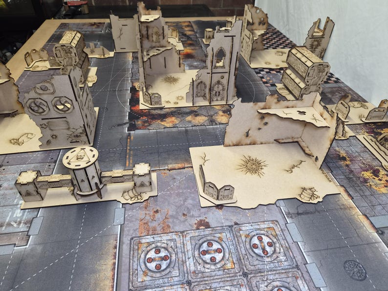 May include: A collection of laser-cut wooden tabletop wargaming terrain pieces arranged on a printed game mat. The set includes various ruined building structures, walls, and defensive barricades featuring etched details and weathered textures.
