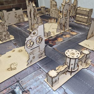 May include: A collection of laser-cut wooden miniature terrain pieces for tabletop gaming, featuring ruined building walls, industrial structures, and cylindrical towers with etched details, arranged on a printed game mat surface.