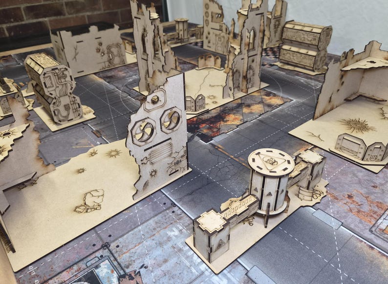 May include: A collection of laser-cut wooden miniature terrain pieces for tabletop gaming, featuring ruined building walls, industrial structures, and cylindrical towers with etched details, arranged on a printed game mat surface.