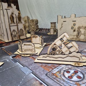 May include: A collection of laser-cut wooden miniature terrain pieces for tabletop gaming, featuring ruined building walls, gothic archways, and barricades with etched details, arranged on a printed game mat with a post-apocalyptic urban design.