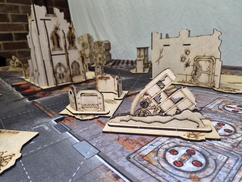 May include: A collection of laser-cut wooden miniature terrain pieces for tabletop gaming, featuring ruined building walls, gothic archways, and barricades with etched details, arranged on a printed game mat with a post-apocalyptic urban design.