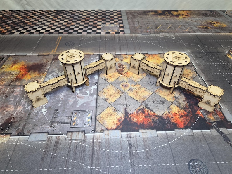May include: A laser-cut wooden tabletop gaming terrain set featuring two cylindrical towers connected by modular bridge sections. The structures are made of light-colored plywood with engraved geometric details and triangular cutouts on the walls.
