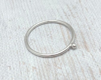 May include: A minimalist silver ring with a small, round bead detail. The ring is thin and delicate, with a simple, modern design. The ring is shown on a light-colored, textured surface.