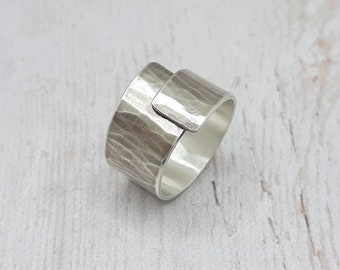 May include: A silver ring with a textured, hammered finish. The ring features a wide band with a rectangular overlay, creating a modern, geometric design. The metal has a slightly matte appearance.