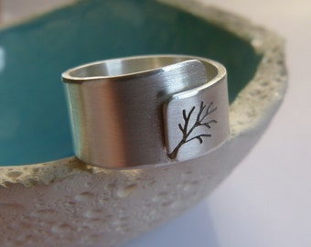 May include: A silver-colored ring with a brushed finish. The ring features a cut-out tree design on a rectangular plate. The ring is wrapped around the finger. The ring is sitting in a light blue bowl.