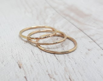 May include: Three delicate gold rings with textured and smooth bands are displayed. The rings are stacked together, showcasing variations in surface design. The rings are made of a shiny gold material.