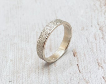 May include: A textured silver ring with a bark-like pattern. The ring has a flat profile and a polished interior. The ring is a simple band design, suitable for everyday wear. The ring is shown on a light-colored wooden surface.
