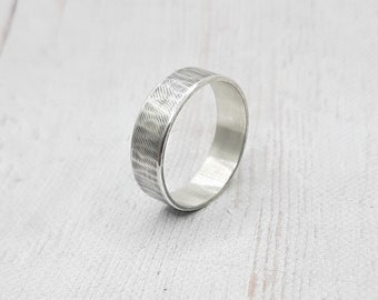 May include: A silver ring with a textured surface. The ring has a band with a pattern of fine, parallel lines. The ring is made of a shiny, metallic material and is displayed on a light-colored surface.