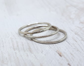 May include: Three silver rings with textured and smooth bands are stacked on a light-colored surface. The rings are circular and appear to be made of metal. The bands have different textures, with one having a hammered look.