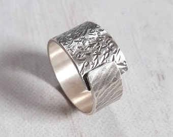 May include: A wide, textured silver ring. The ring features a detailed, embossed design on the top and a contrasting linear pattern on the side. The metal has a bright, polished finish, creating a striking contrast with the textured surfaces. The ring is a piece of jewelry.