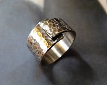 May include: A silver ring with a hammered texture on the top band. The ring has a unique design with a small, overlapping section. The metal has a shiny, reflective quality, and the ring appears to be handmade.