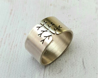 May include: A silver-colored ring with a tree branch design. The ring is wide and has a brushed metal finish. The tree branch design is cut out of the metal, creating a negative space effect. The ring is on a white surface.