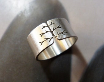 May include: A silver-colored ring with a tree branch design. The ring is wide and has a brushed metal finish. The tree branches are cut out of the metal, creating a negative space design.
