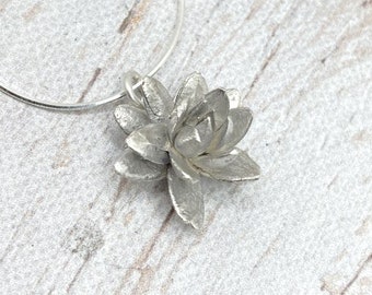 May include: A silver-colored pendant necklace featuring a detailed, three-dimensional flower design. The flower has multiple petals and is suspended from a thin, silver-colored chain. The pendant is likely made of metal and has a textured surface.