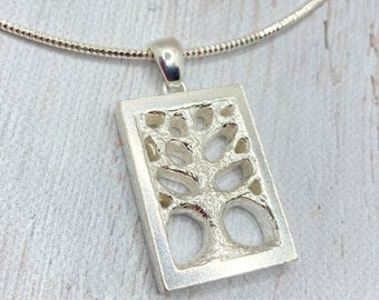 May include: A silver pendant necklace featuring a rectangular pendant with a tree design cut out. The pendant is suspended from a thin, silver chain. The tree design has a textured appearance, and the necklace is displayed on a light-colored surface.