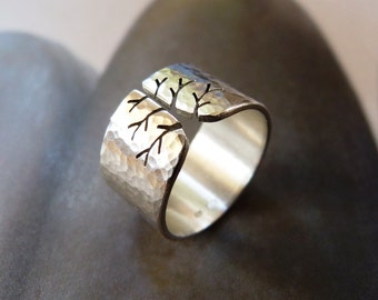 May include: A silver-colored ring with a hammered texture and a tree branch design. The ring has a split design with the tree branches etched on the top. The ring is a wide band style.