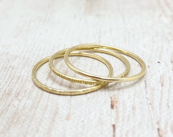 May include: Three delicate gold rings. One ring has a hammered texture, another has a linear pattern, and the third is smooth. The rings are stacked together, creating a minimalist and elegant jewelry set.