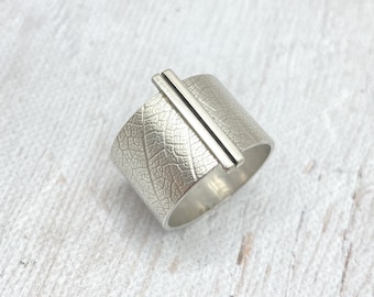May include: A silver ring with a wide band featuring a leaf texture. A rectangular bar is set across the top of the ring. The ring has a nature-inspired design and is likely made of sterling silver.