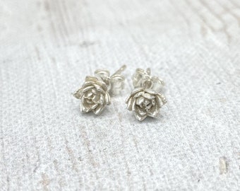 May include: A pair of small, silver-colored stud earrings. Each earring is shaped like a detailed succulent flower. The earrings are displayed on a textured, light-colored surface, showcasing their intricate design and metallic sheen.