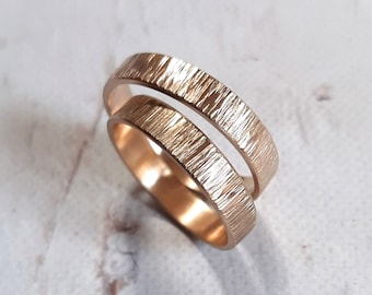 May include: Two rose gold wedding bands with a textured, wood-grain-like surface. The rings are stacked, showcasing their similar design and warm metallic color. The bands are set against a neutral, slightly mottled background.