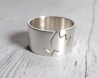 May include: A wide silver band ring featuring a brushed matte finish and a single engraved tulip outline. The design includes a vertical cut that separates the flower motif from the rest of the band, creating a clean and simple aesthetic.
