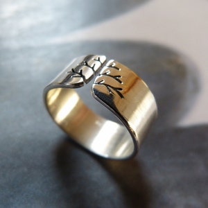 Op de afbeelding: Een zilverkleurige ring met een gespleten ontwerp. De ring heeft een geborsteld gouden binnenkant en een gedetailleerd boomtakpatroon op de buitenranden. Het open ontwerp maakt een verstelbare maat mogelijk.