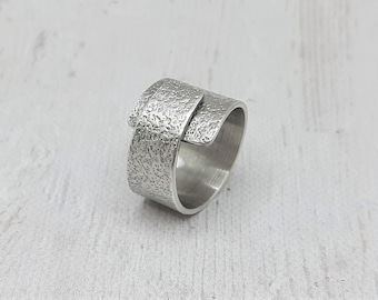 May include: A textured silver ring with an overlapping design. The ring has a wide band with a rough, hammered appearance. The top section of the ring overlaps the band, creating a unique, modern look. The ring is shown on a light-colored surface.
