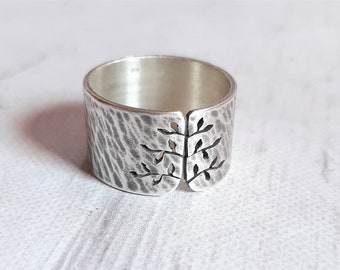 May include: A wide, silver ring with a textured surface and a cut-out tree design. The ring has a hammered appearance and a split design at the front, showcasing the tree motif. The ring is a piece of jewelry.