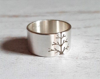 May include: A silver ring with a brushed finish. The ring features a cut-out tree design on the band. The ring is a wide band style, with a simple and elegant aesthetic. The ring is likely made of sterling silver.