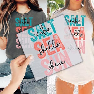 May include: Two t-shirts, one dark gray and one cream, each with the words "water, sea, shells, sun, shine" in a layered design. The text is in teal, coral, and pink, with a handwritten font.