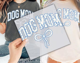 May include: A white t-shirt with a blue and white patterned design that says "DOG MOM" with a blue bow underneath. The shirt is being held by a person wearing a gray t-shirt with the same design.