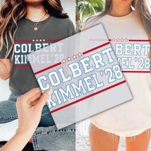 May include: Two t-shirts, one dark gray and one cream, each displaying the text "COLBERT KIMMEL '28" in white, outlined in light blue, with red stripes and star accents. The shirts appear to be casual wear.
