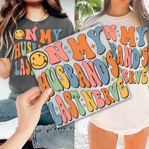 May include: Two t-shirts with the text "ON MY HUSBAND'S LAST NERVE" in colorful, retro-style lettering. One shirt is dark gray, the other is off-white. The design includes a smiley face and star accents.