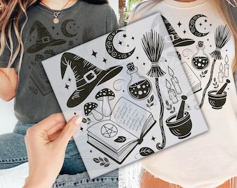Puede incluir: Una camiseta de manga corta con un estampado gráfico negro de un sombrero de bruja, una luna creciente, estrellas, setas, una poción, una escoba, velas, un mortero con mano y un libro de hechizos abierto con un símbolo de pentáculo.